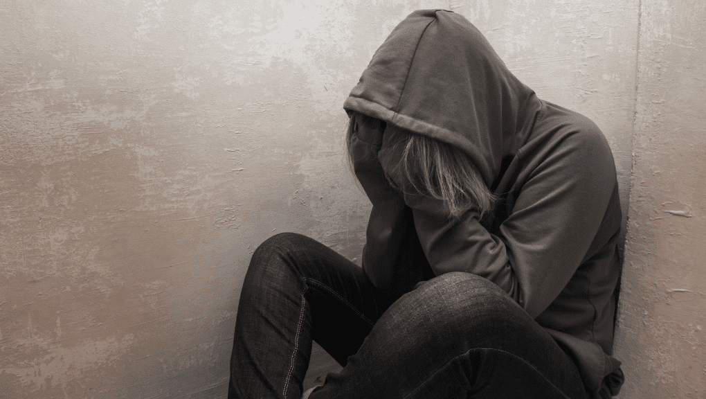 A person wearing a hoodie sits on the floor, leaning against a plain wall and covering their face with their hands, appearing distressed or upset. The surroundings are minimal, and the lighting is subdued, contributing to a somber mood.